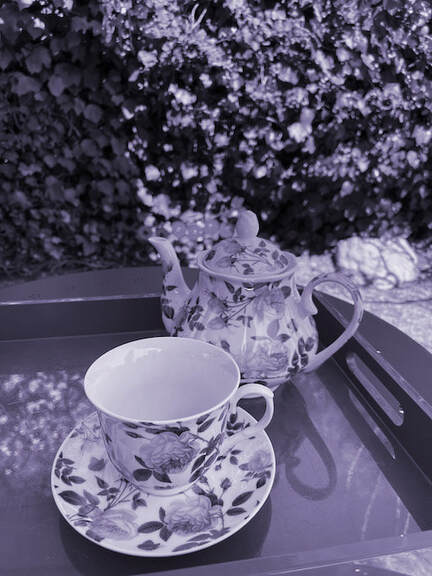 In a light purple haze, beautiful leafed china tea cup and saucer and teapot in a rectangular wooden tray sitting on a small round table outside in a garden with a full hedge behind.